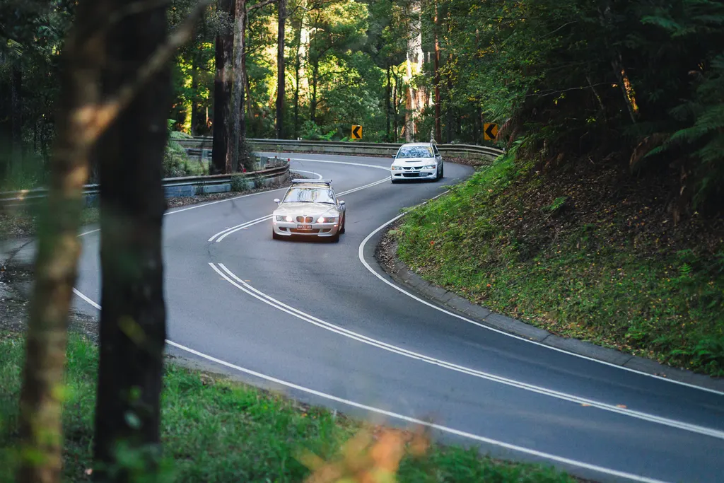 Two cars navigate a winding forest road with protective barriers and curve warning signs.