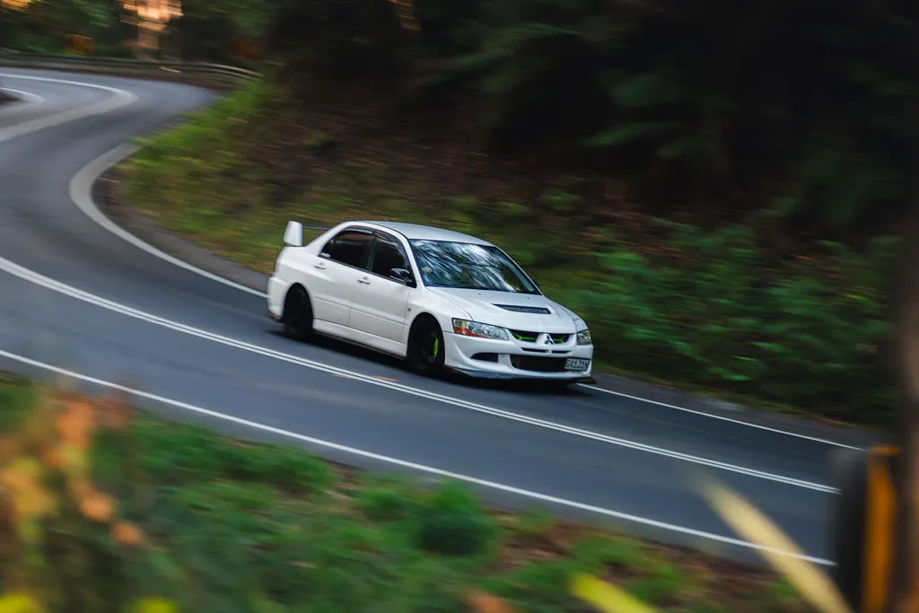 White sedan driving through a curve on a mountain road surrounded by green forest.