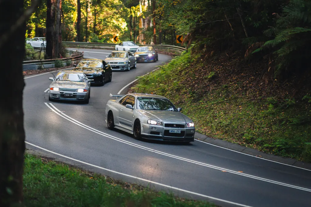 A silver sports car leads a convoy of vehicles through a forested mountain road lined with tall trees and dense foliage.