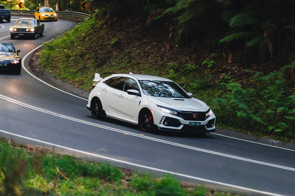 White performance hatchback navigating a forested mountain road with other vehicles visible in the distance.