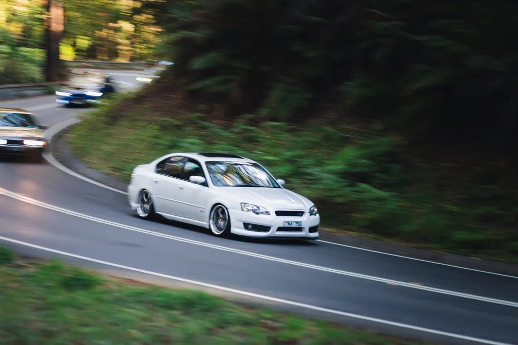 White sedan cornering on a tree-lined road with motion blur in background.