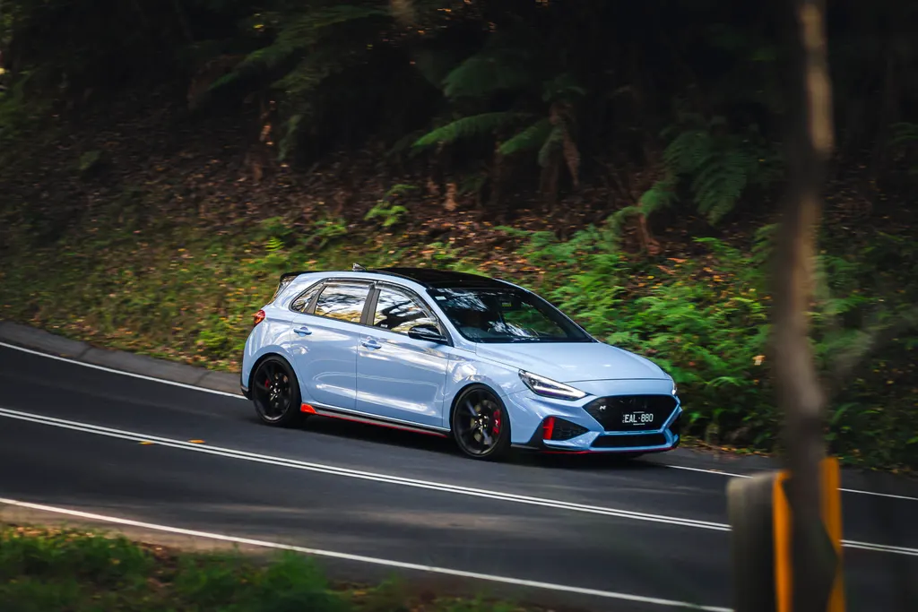Light blue hatchback with red accents on a winding forest road with trees and undergrowth in the background.