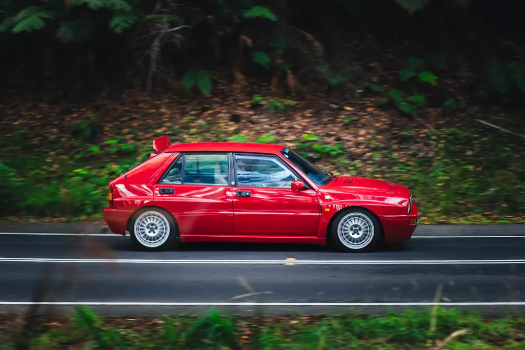 Red compact sedan driving on a two-lane road bordered by dense green forest.