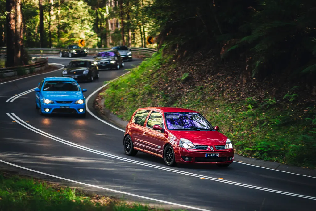 A red hatchback leads a blue sedan through a winding forest road lined with tall trees and moss-covered embankments.