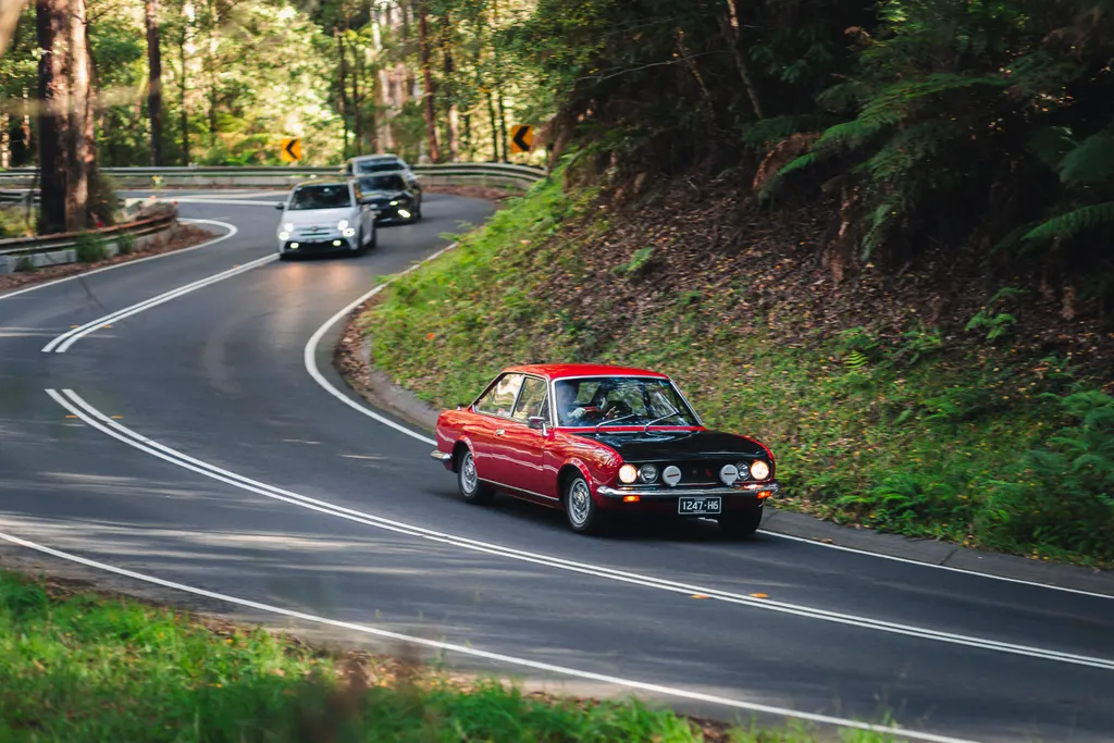 A red vintage saloon car leads a group of vehicles through a winding forest road with dense green vegetation and tall trees lining both sides.