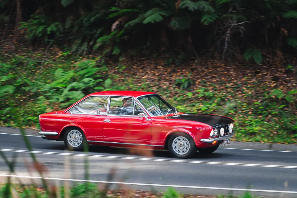 Red vintage coupe with auxiliary lights driving along a forest road lined with dense green foliage.
