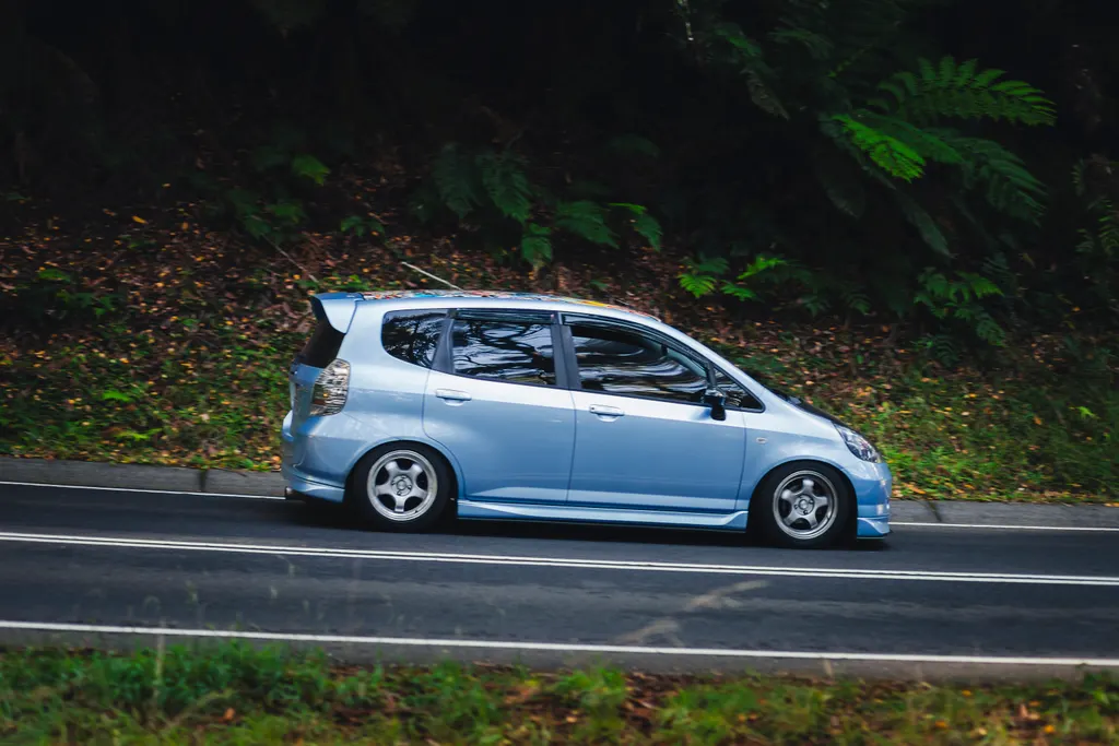 Light blue modified compact hatchback driving on a road with dense forest foliage in the background.
