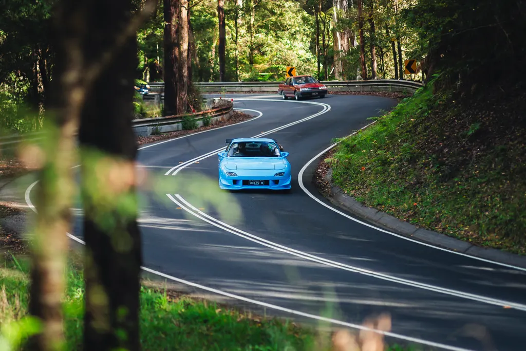 A blue sports car navigates a winding forest road with tall trees lining both sides, while a red vehicle follows in the distance.