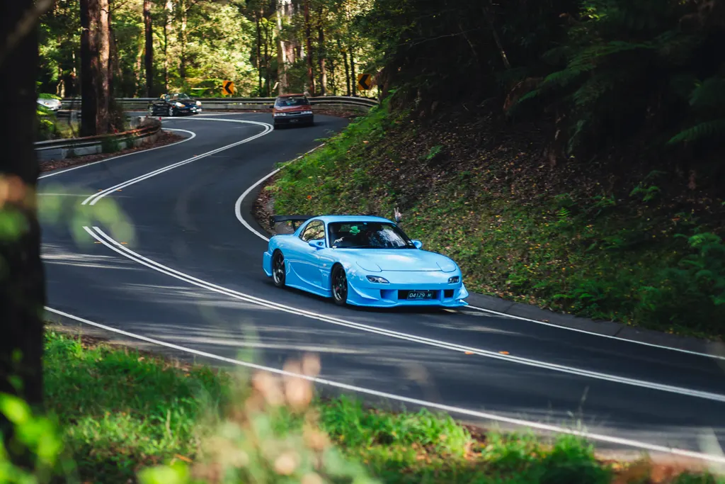 Blue sports car navigating a winding forest road with other vehicles following behind.