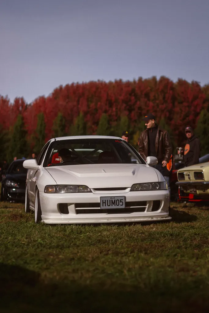 White modified sedan displayed at outdoor car show with spectators and coniferous trees in background.