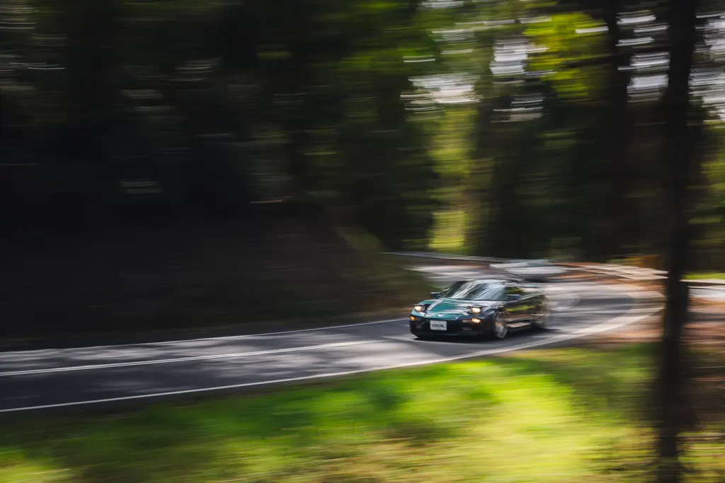 A teal-colored coupe drives through a tree-lined road with motion blur surrounding the vehicle and forest.