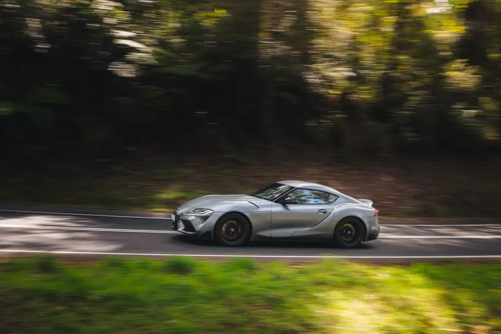 A silver sports car with gold wheels drives along a tree-lined road with motion blur surrounding the vehicle.