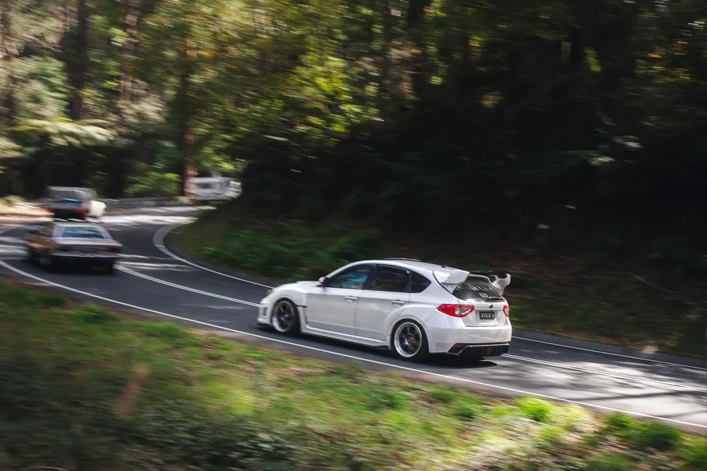 White performance hatchback driving on a curved road through dense forest with other vehicles visible ahead.