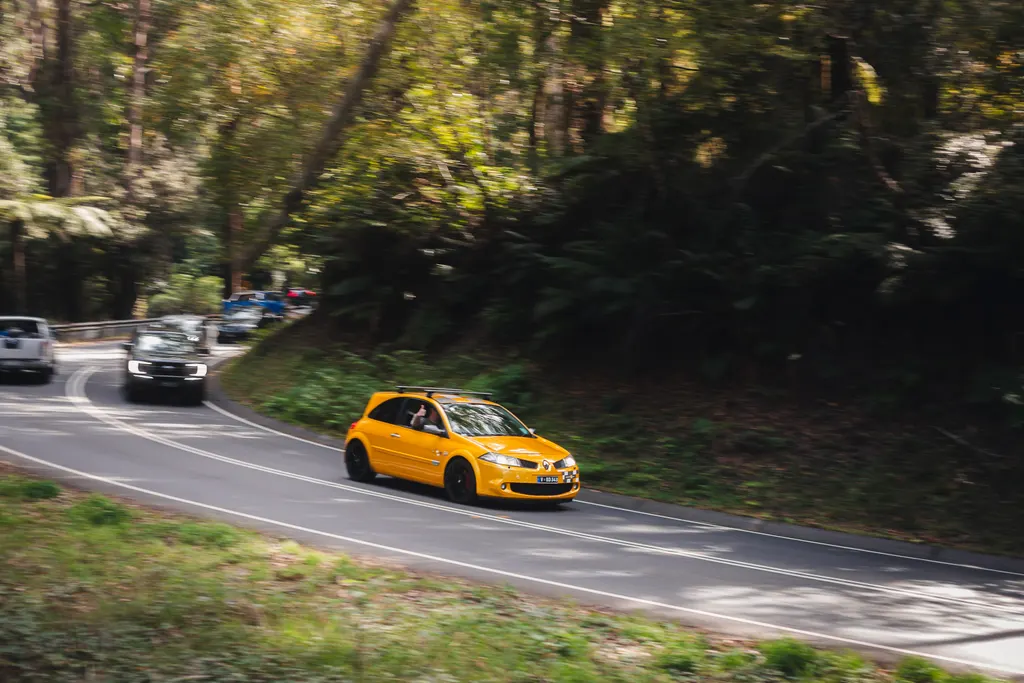 Yellow sports car navigating a tree-lined road curve with other vehicles visible ahead.