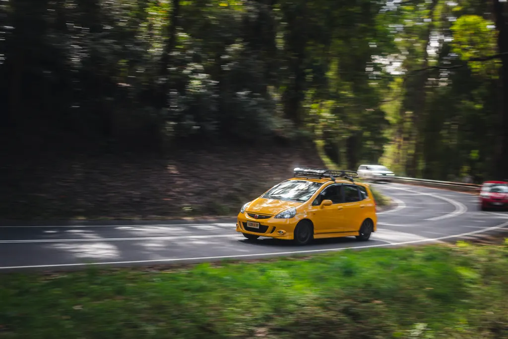 Yellow hatchback navigating a forest road curve with motion blur surrounding foliage.
