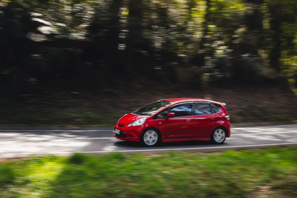 Red hatchback car driving on a tree-lined road with motion blur in the background.