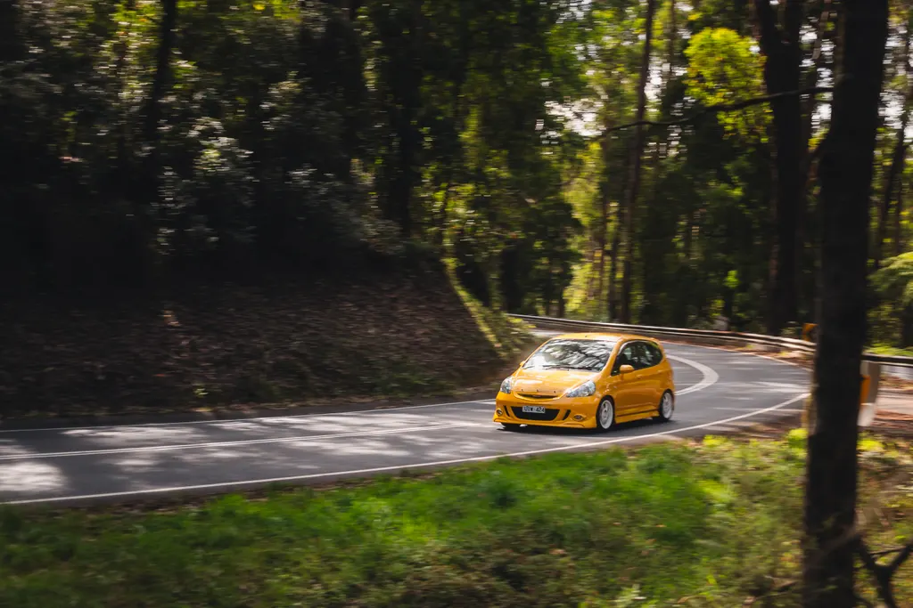 Yellow sports hatchback navigating a forest road with trees lining both sides.