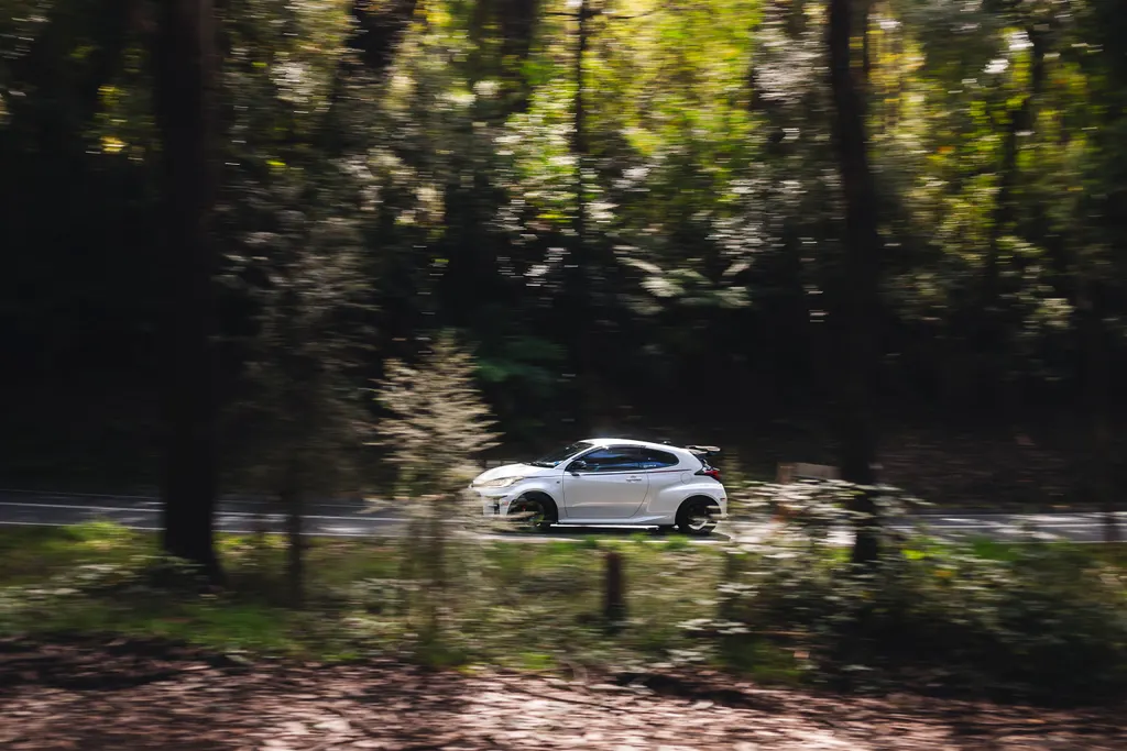 White compact car driving through a tree-lined forest road with motion blur surrounding the vehicle.