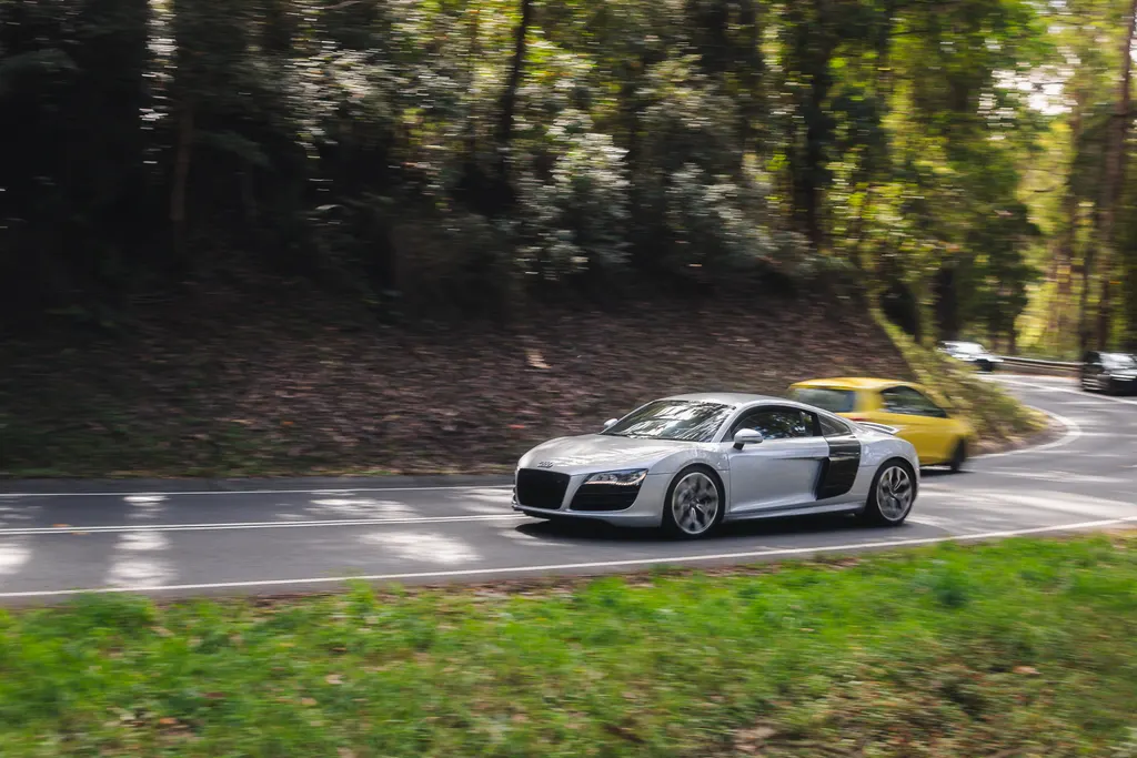 Silver sports car with open door driving on a tree-lined road with a yellow car visible behind.