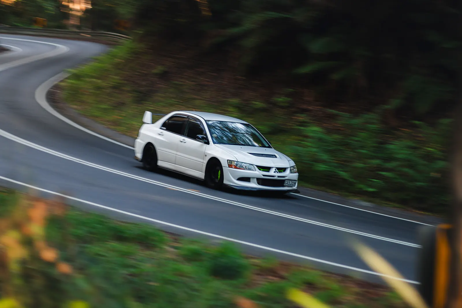 White sedan driving through a curve on a mountain road surrounded by green forest.