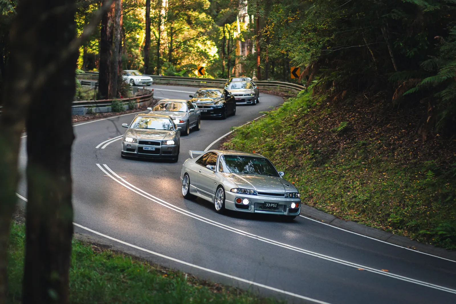 A convoy of Japanese sports cars navigates a winding forest road lined with tall trees and lush green vegetation.