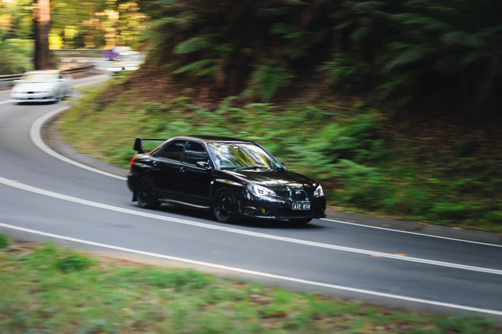 Black sedan navigating a curved road through dense green forest with a second vehicle visible on the road behind it.