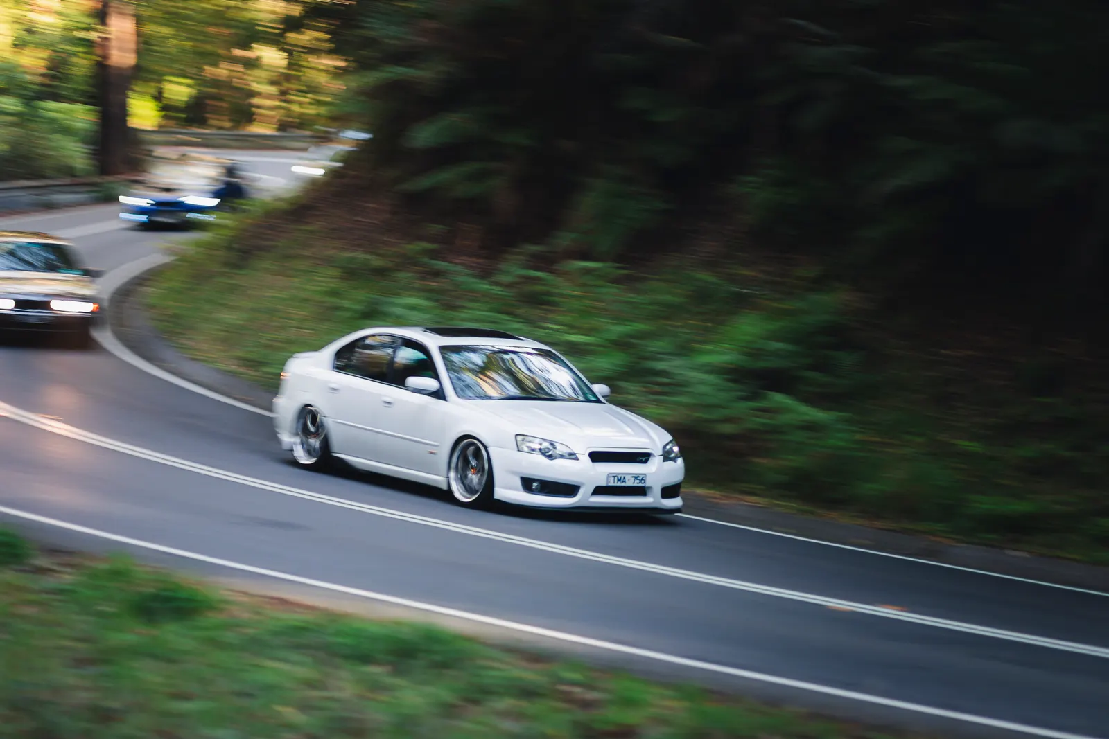 White sedan cornering on a tree-lined road with motion blur in background.