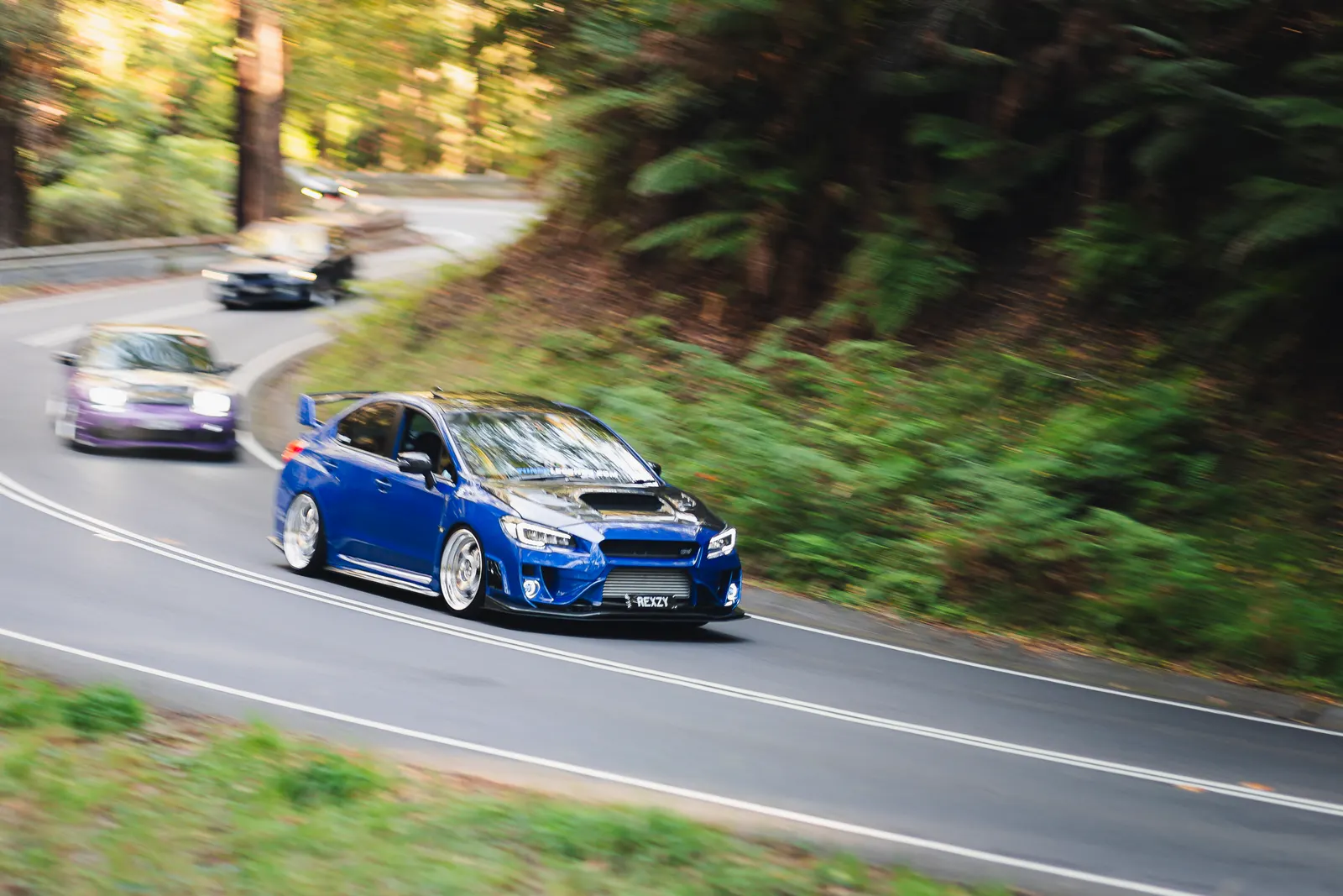 Blue sports sedan leading a convoy of vehicles through a wooded mountain road with motion blur.