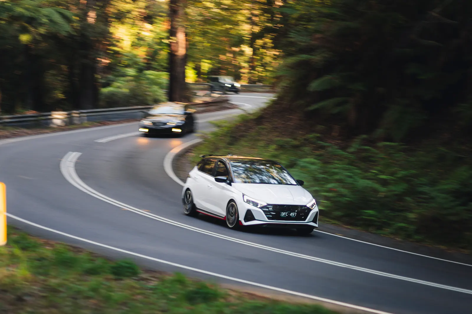 White sports car leading a convoy through a tree-lined road with trailing vehicles visible behind on a curved bend.