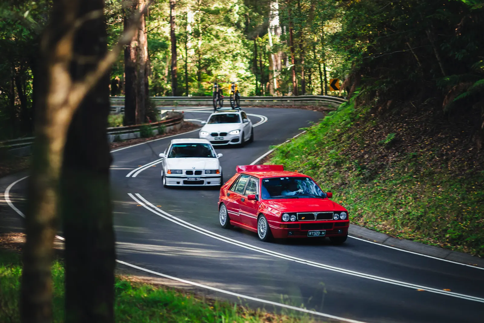 Three performance cars navigate a tree-lined mountain road in single file, with a red compact sports car leading two white sedans through a sweeping curve.