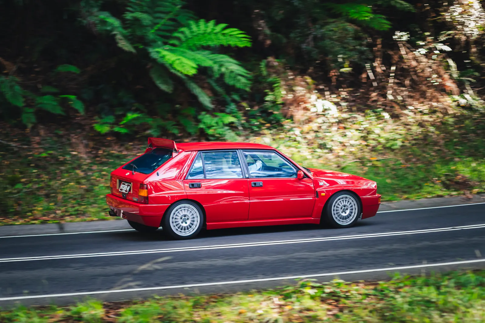 Red sedan driving on a forest road with green foliage on both sides.
