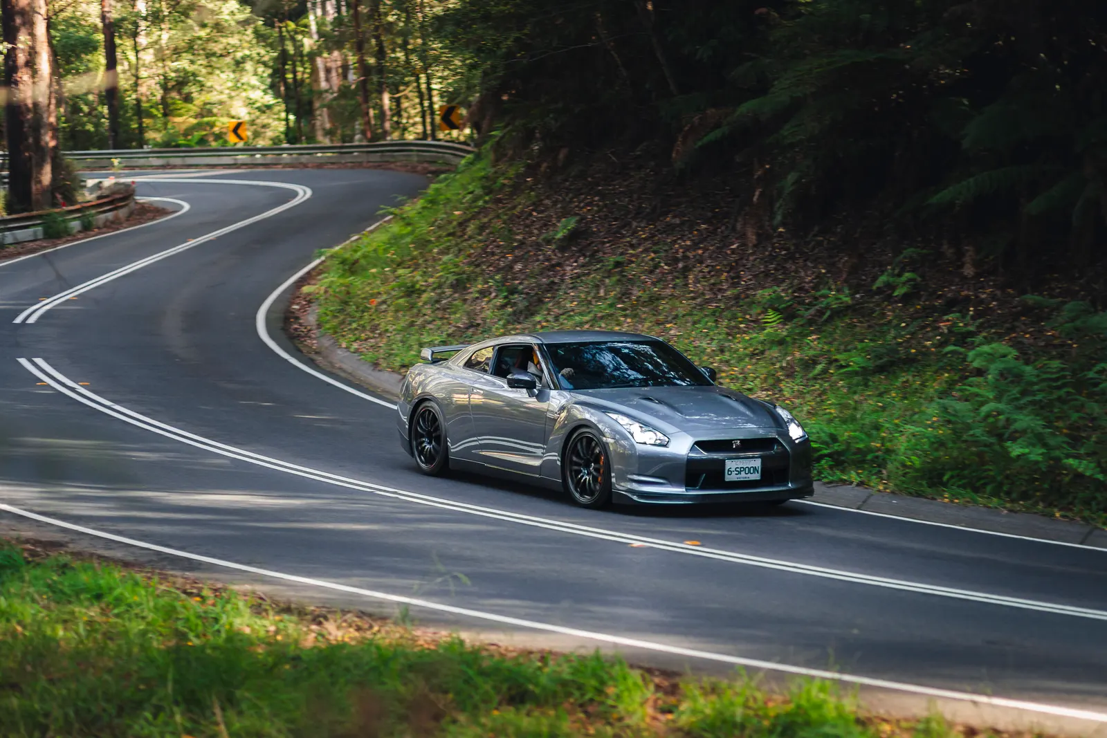 Silver sports car navigating a curved forest road with dense green vegetation on both sides.