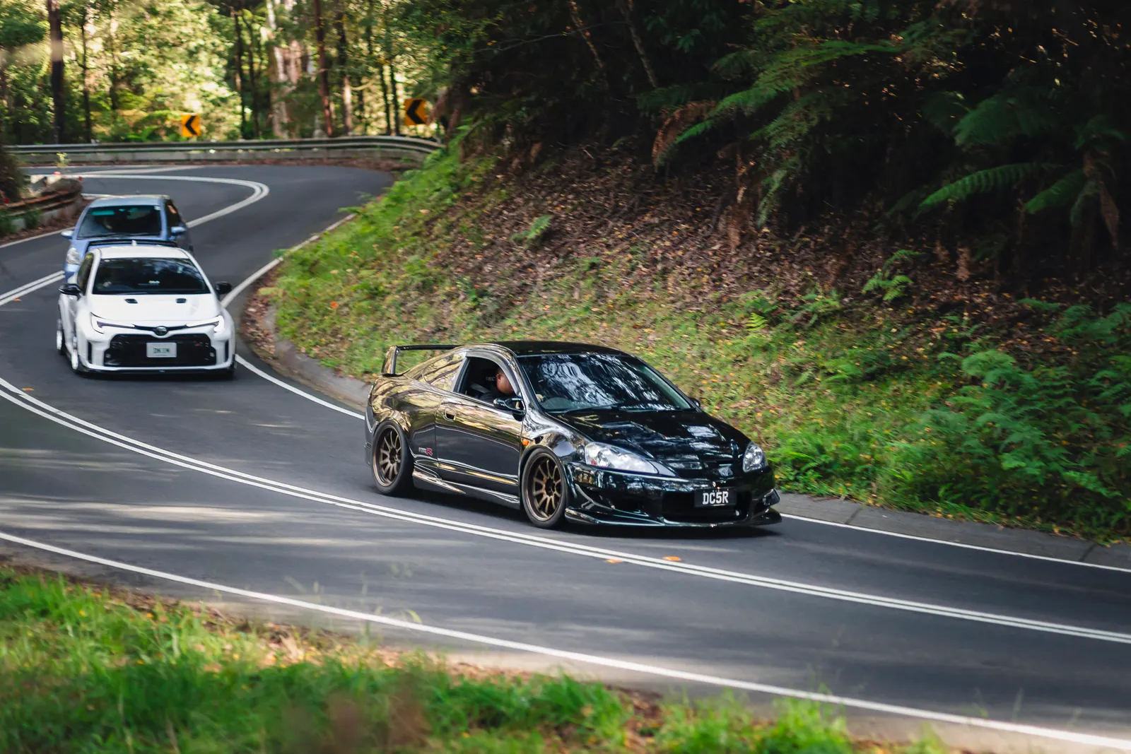 A black sports sedan leads two white vehicles through a curved forest road lined with dense green foliage.