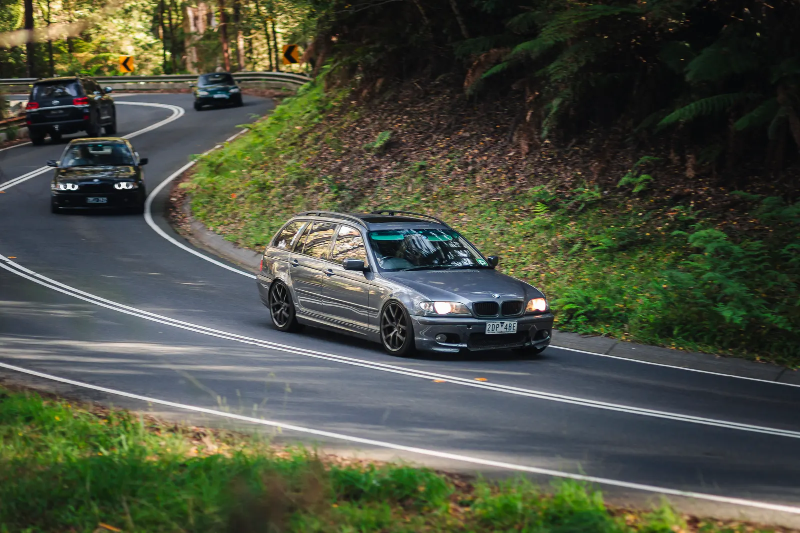 Silver sedan leading a group of cars through a wooded mountain road with tight curves and lush green vegetation.