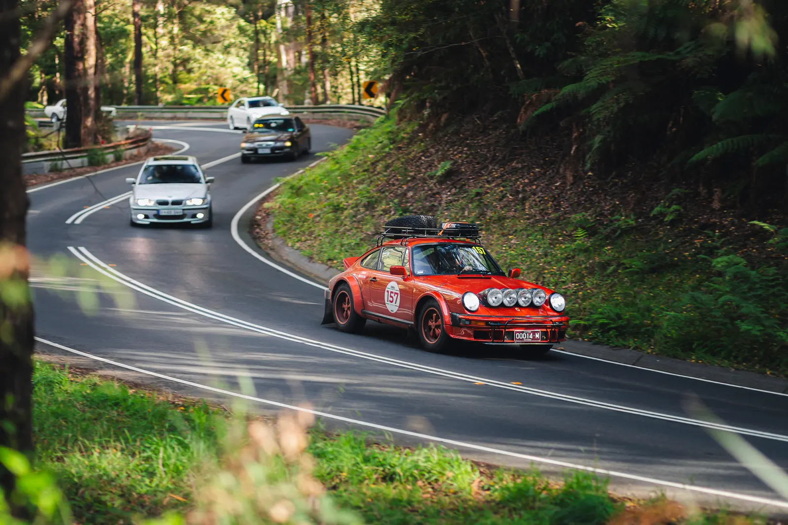 A red rally car with roof-mounted lights leads a convoy of vehicles through a forested mountain road with tight curves.