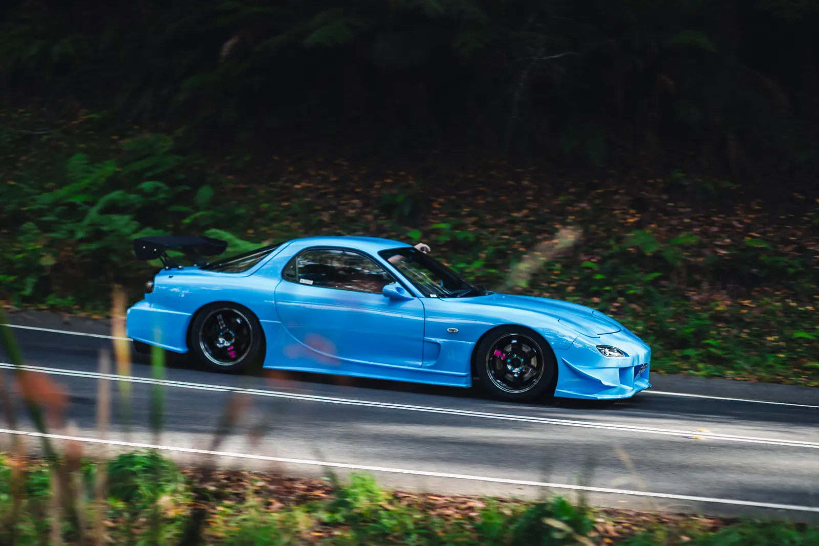 Bright blue sports car with aggressive aerodynamic body kit on a wet road bordered by green forest.