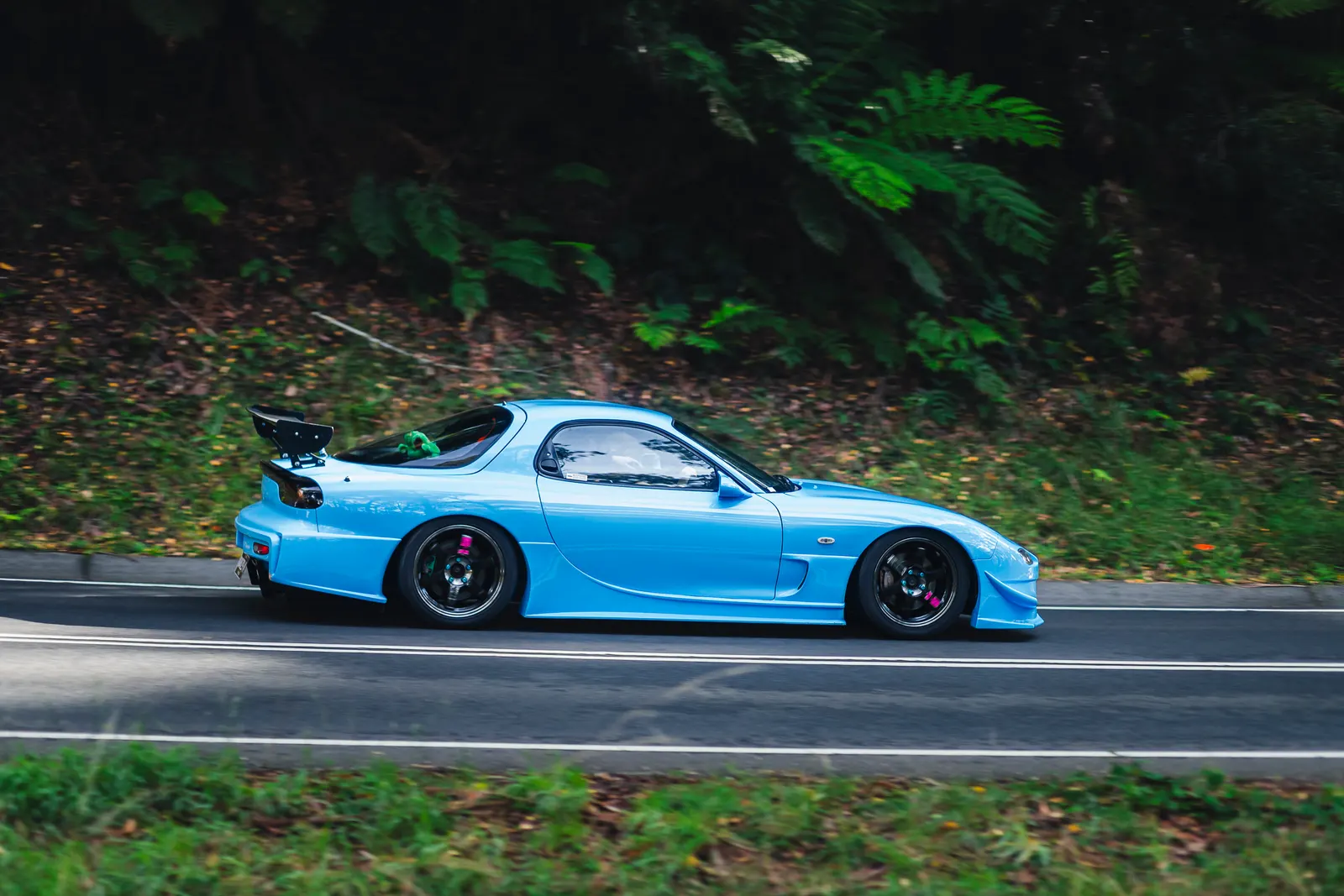 Blue sports car with large rear wing on a forest road, captured in profile.