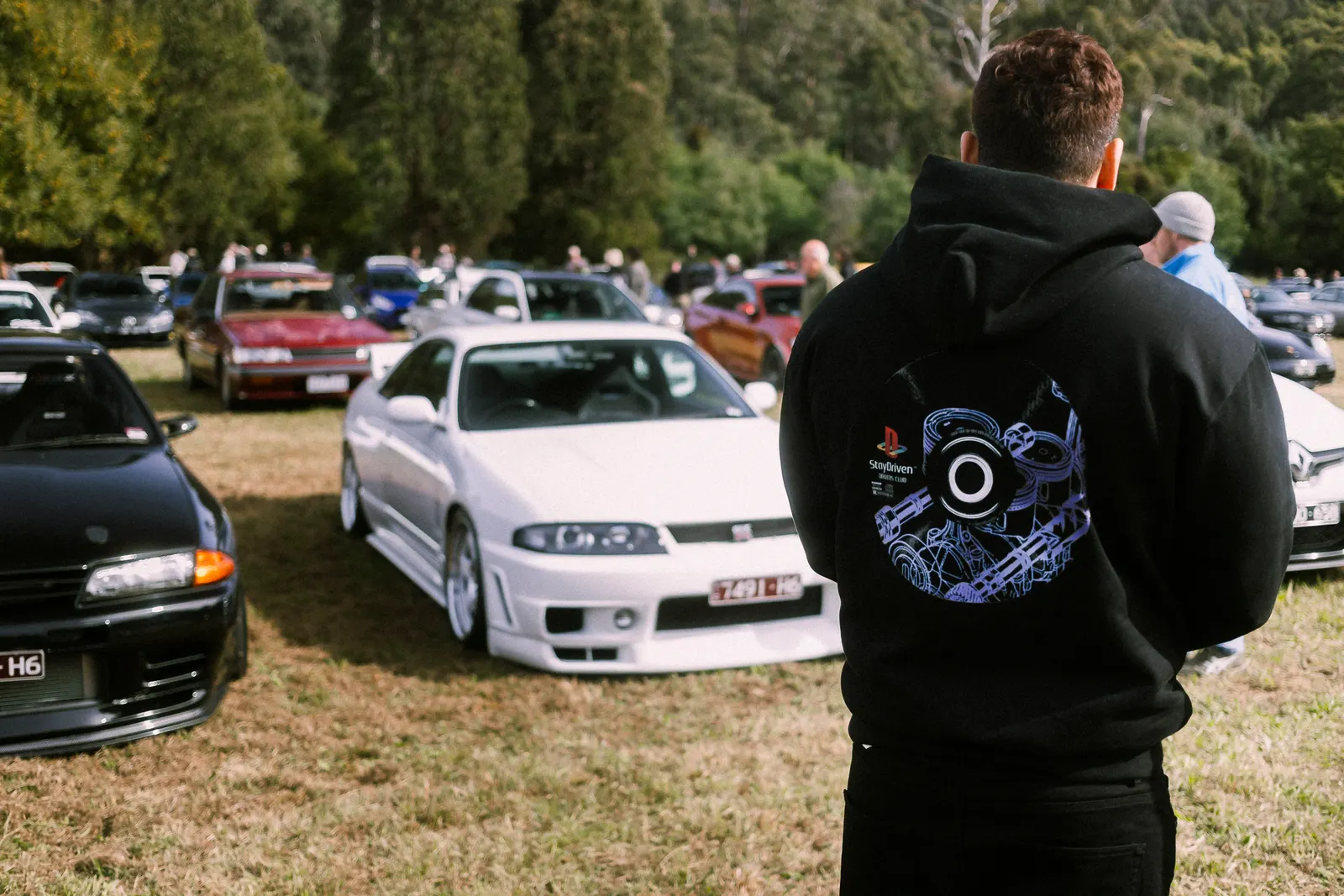 A person in a black hoodie stands in a grass field viewing parked classic cars at an outdoor automotive gathering.