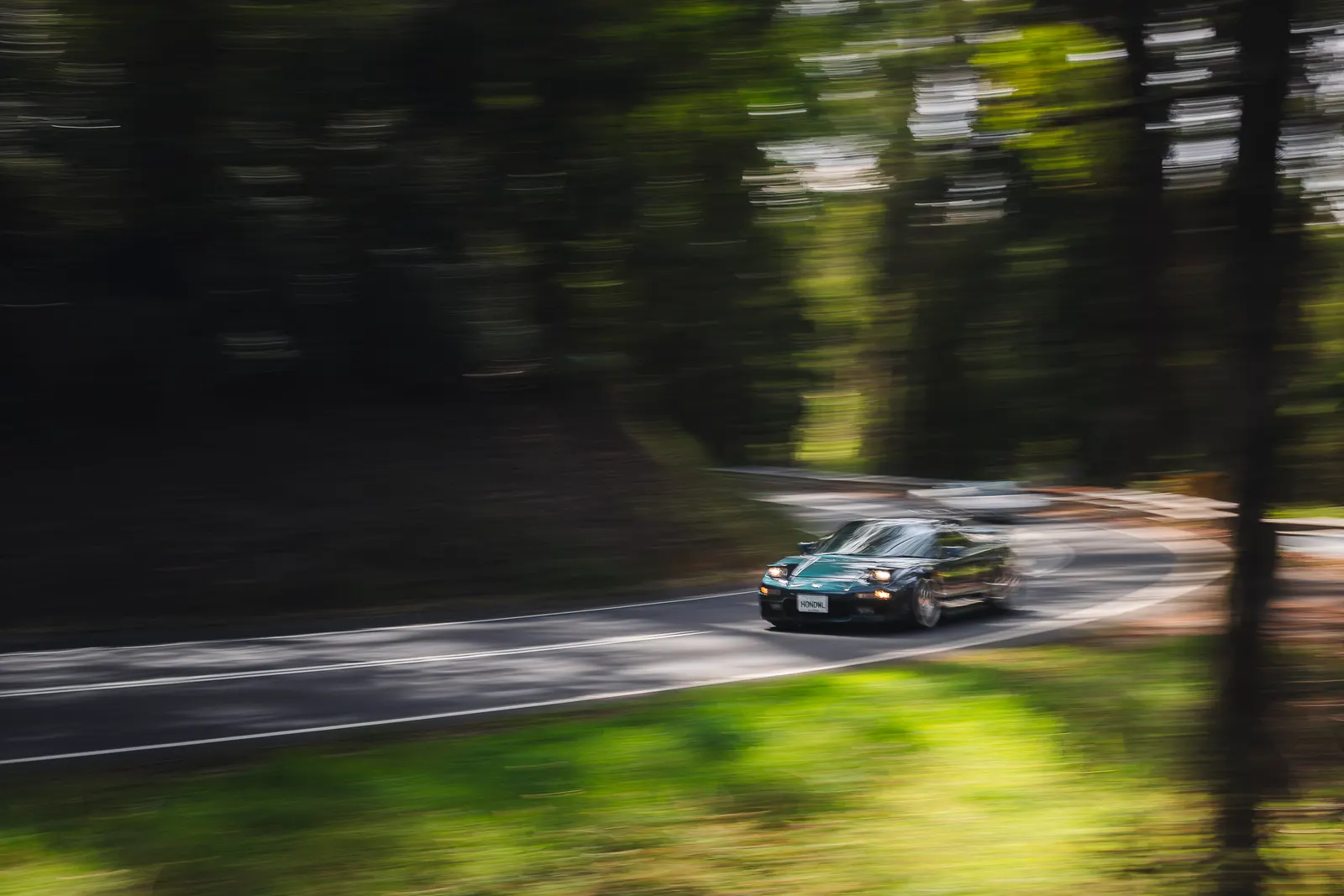 A teal-colored coupe drives through a tree-lined road with motion blur surrounding the vehicle and forest.