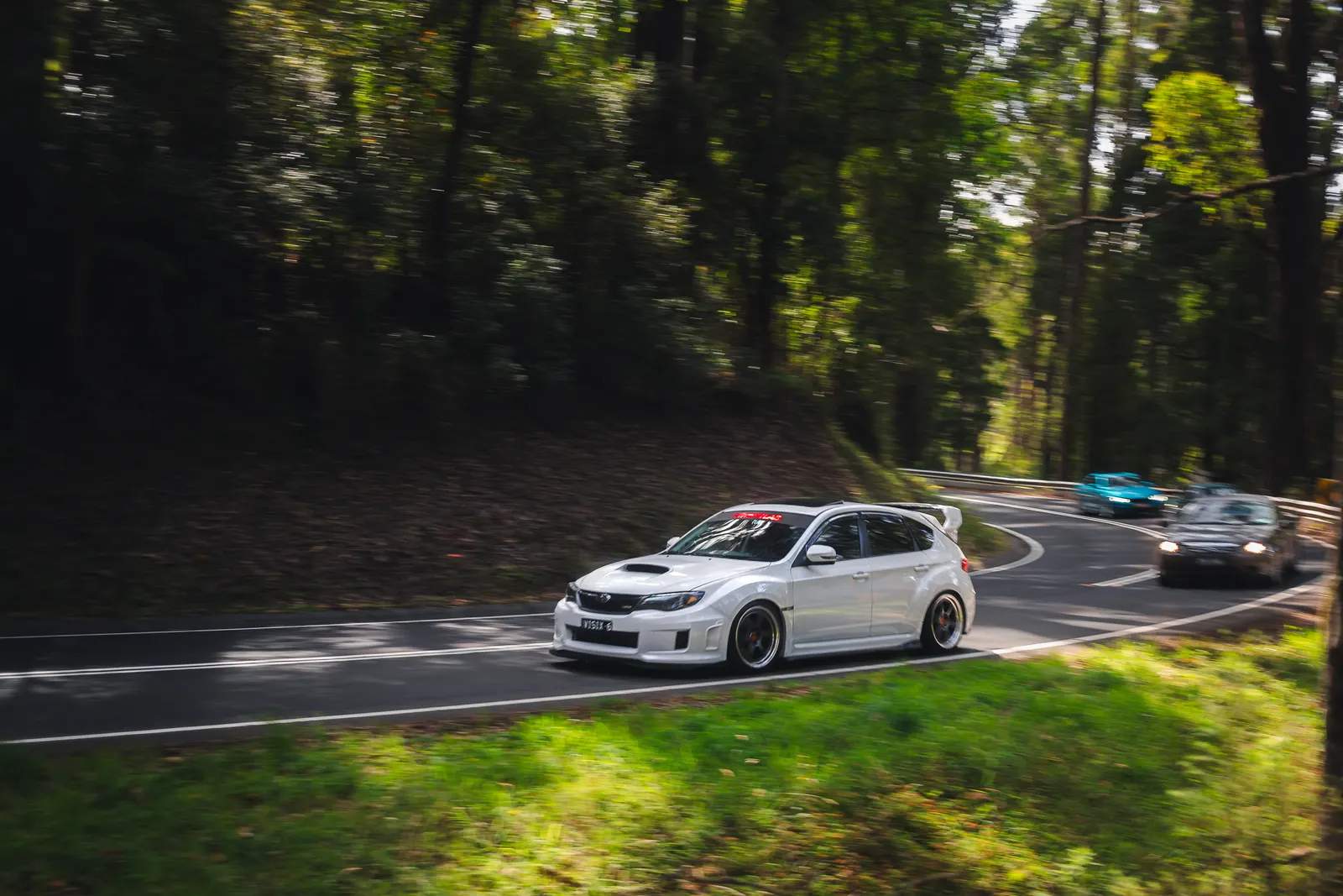 White sport hatchback cornering on a tree-lined road with other vehicles visible in the background.