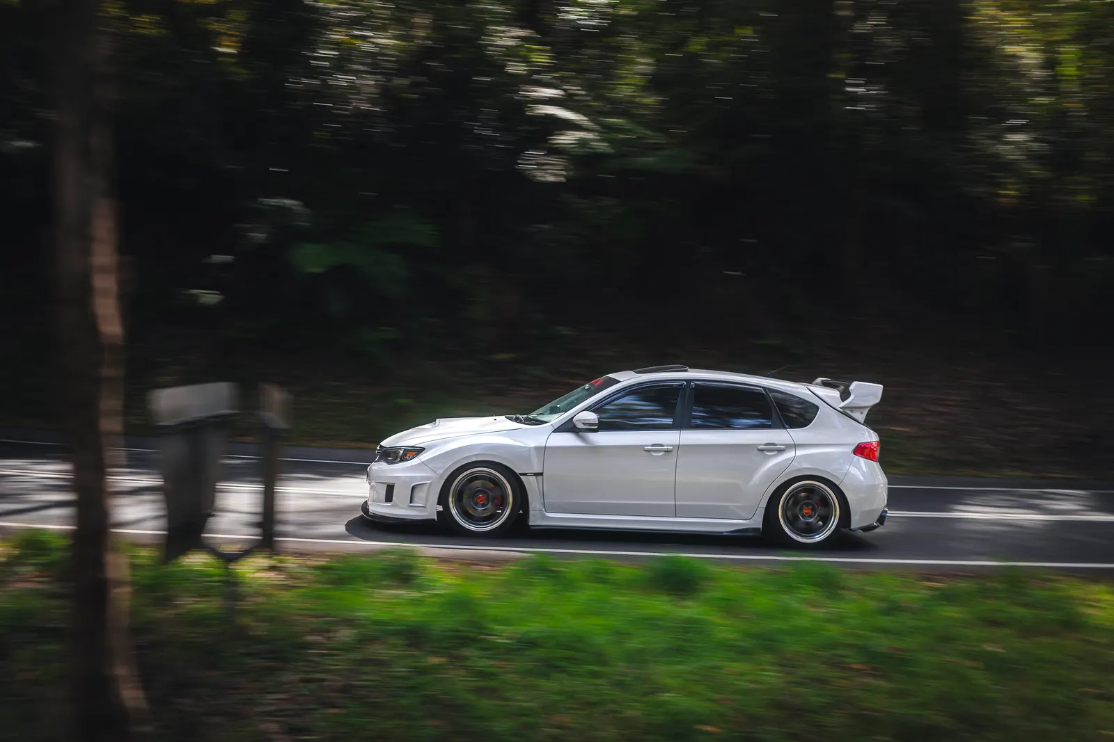 White compact hatchback with aggressive bodywork and red brake calipers driving on a tree-lined road.