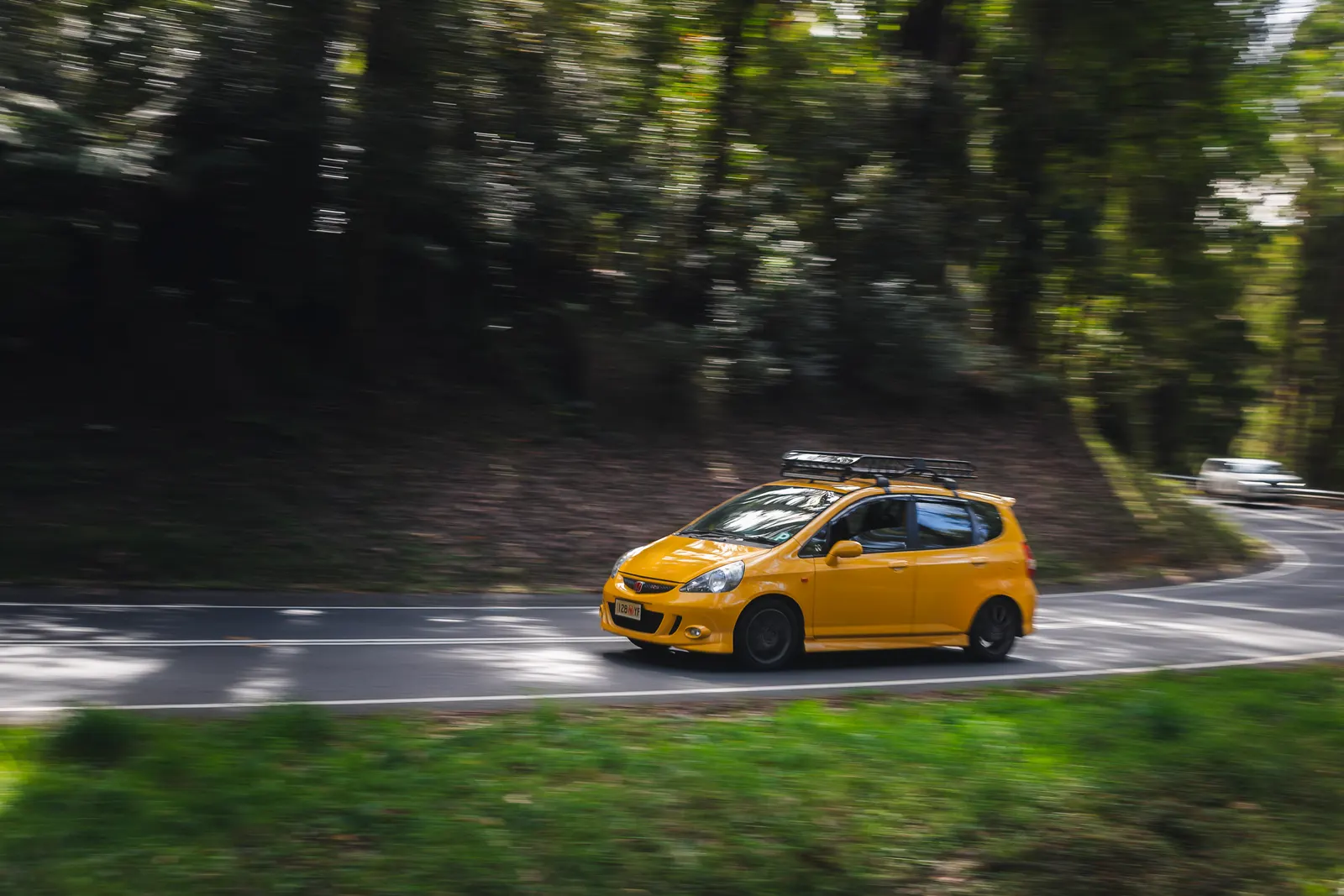 Yellow hatchback car driving through a tree-lined road with motion blur in the background.