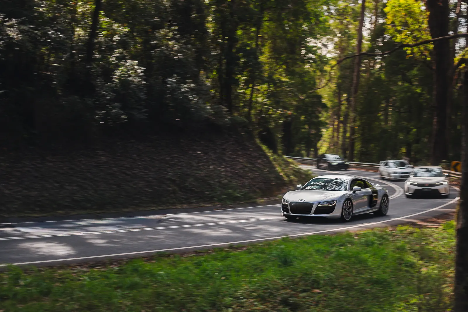 A silver Audi supercar leads a convoy of vehicles through a forested road with dense green trees lining both sides.