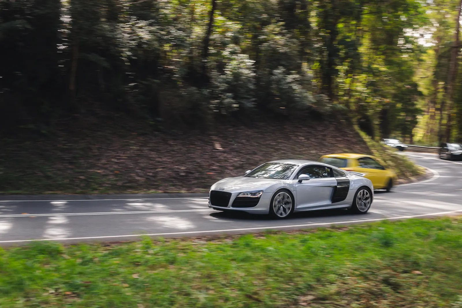Silver sports car with open door driving on a tree-lined road with a yellow car visible behind.