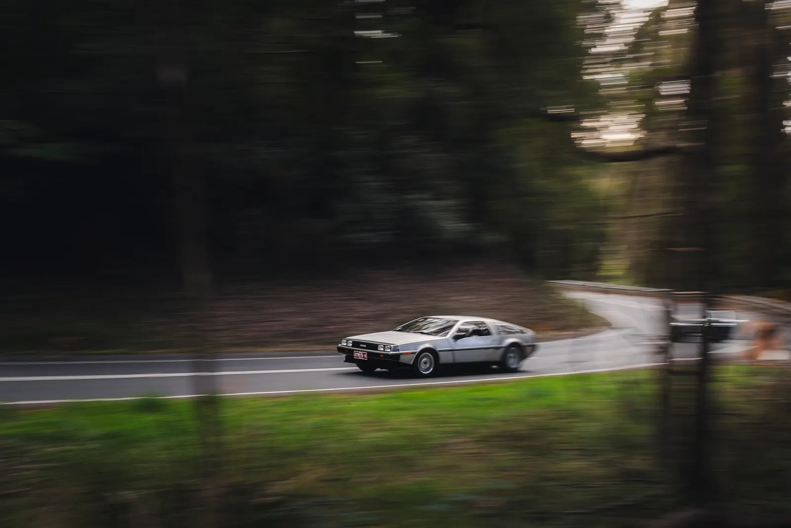 A silver DeLorean sports car driving on a road lined with trees, captured with motion blur.
